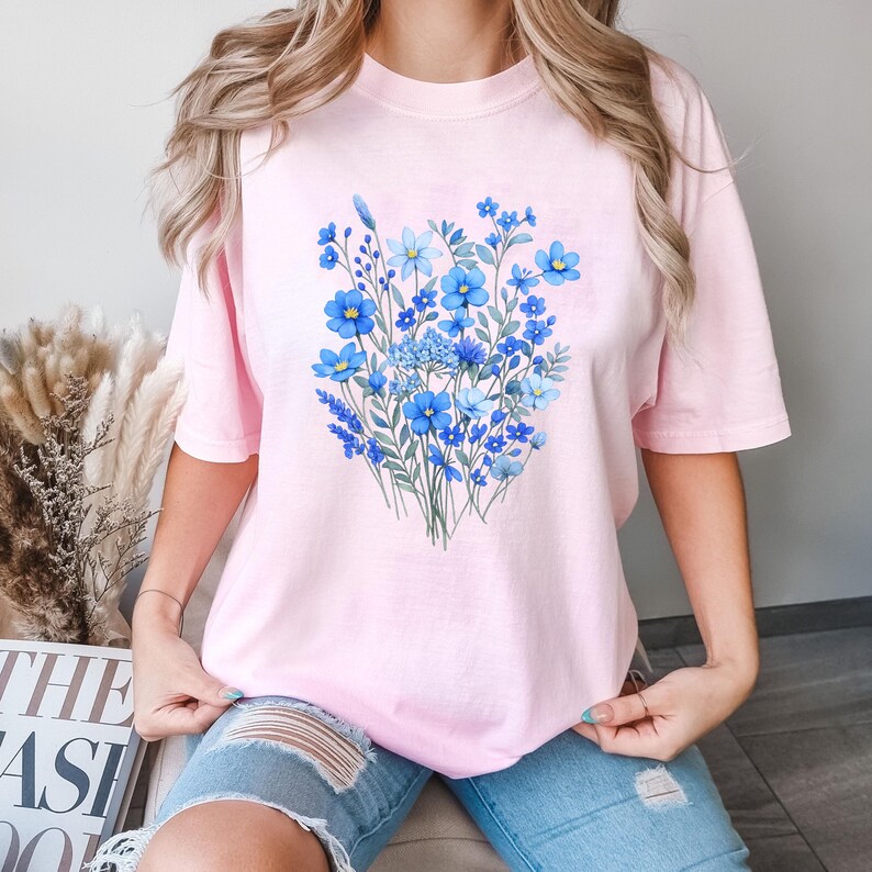 May include: A light pink t-shirt featuring a vibrant bouquet of blue flowers. The floral design includes various shades of blue, with green stems and leaves. The shirt is a soft, casual style.
