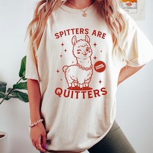 May include: A cream-coloured t-shirt featuring a red graphic of a winking llama wearing a floral necklace. The text "SPITTERS ARE QUITTERS" is above and below the llama. A small badge reads "WINNERS SWALLOW".