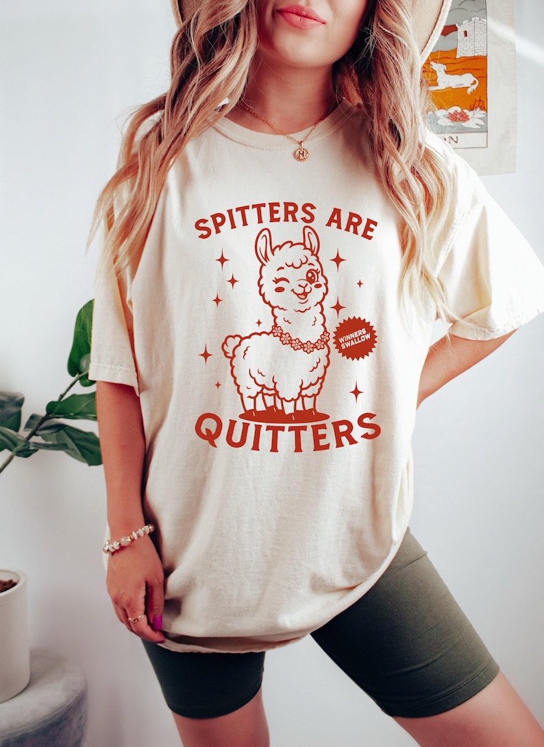 May include: A cream-coloured t-shirt featuring a red graphic of a winking llama wearing a floral necklace. The text "SPITTERS ARE QUITTERS" is above and below the llama. A small badge reads "WINNERS SWALLOW".