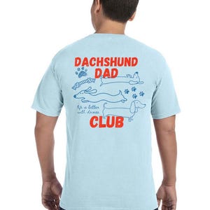 May include: Pale blue t-shirt with the words "DACHSHUND DAD CLUB" in red and blue. The shirt features a graphic of dachshunds and paw prints. The word "CHAMBRAY" is printed at the bottom.