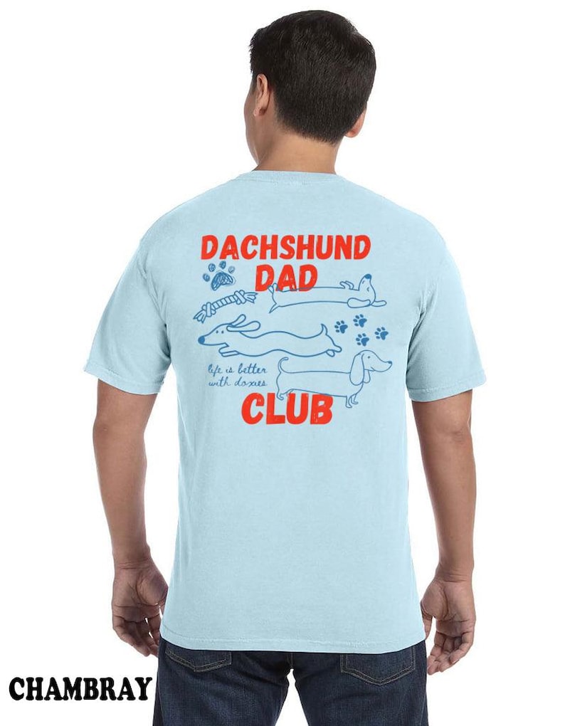 May include: Pale blue t-shirt with the words "DACHSHUND DAD CLUB" in red and blue. The shirt features a graphic of dachshunds and paw prints. The word "CHAMBRAY" is printed at the bottom.