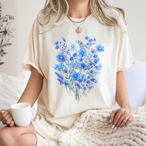 May include: Cream-colored t-shirt featuring a watercolor floral design in shades of blue. The design includes various flowers and foliage. The shirt is a relaxed fit and has short sleeves. The person is holding a white mug.