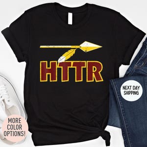 May include: Black t-shirt featuring a distressed arrow graphic with yellow and white accents, and the word "HTTR" in maroon with a yellow outline. The shirt is paired with blue jeans and white sneakers.