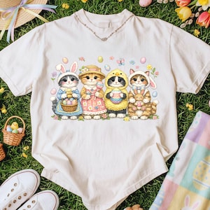 May include: A white t-shirt featuring a colorful illustration of four cats dressed in Easter-themed costumes. The cats are wearing bunny ears, chick costumes, and holding Easter eggs and baskets. The design includes pastel-colored eggs and flowers.
