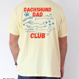 May include: A pale yellow t-shirt with a graphic on the back. The design includes the words "DACHSHUND DAD CLUB" in red, with blue dachshund illustrations and the phrase "life is better with doxies."