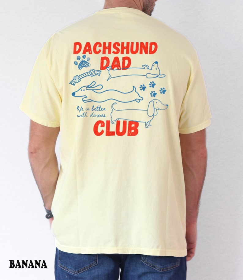 May include: A pale yellow t-shirt with a graphic on the back. The design includes the words "DACHSHUND DAD CLUB" in red, with blue dachshund illustrations and the phrase "life is better with doxies."
