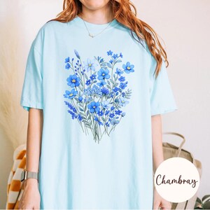 May include: Light blue t-shirt featuring a watercolor floral design in shades of blue. The bouquet includes various flower types and green leaves. The shirt has short sleeves and a relaxed fit, perfect for casual wear.