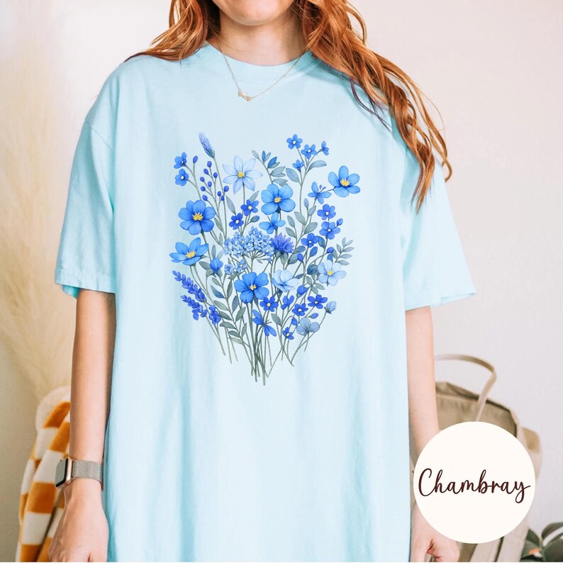 May include: Light blue t-shirt featuring a watercolor floral design in shades of blue. The bouquet includes various flower types and green leaves. The shirt has short sleeves and a relaxed fit, perfect for casual wear.