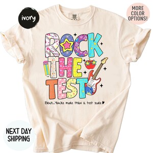 May include: An ivory short-sleeve t-shirt featuring the colorful text "ROCK THE TEST" with graphic elements including a pencil, a red apple with star-shaped glasses, and an electric guitar. Below the text is "Bruh...You're more than a test score".