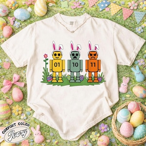 May include: Ivory Comfort Colors t-shirt featuring three robot-like figures with bunny ears, each with a different expression and number. The figures stand on green grass with Easter eggs. The shirt has a V-neck.