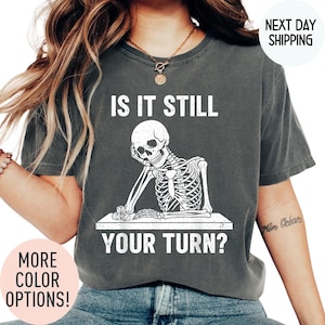 May include: Dark gray t-shirt with the text "IS IT STILL YOUR TURN?" and a white skeleton graphic. The skeleton is leaning on a table with dice. Additional text reads "MORE COLOR OPTIONS!" and "NEXT DAY SHIPPING."