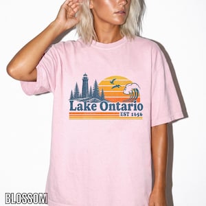 May include: A light pink short-sleeve t-shirt featuring a graphic print of a lighthouse, pine trees, a wave, and birds against a sunset. The text reads Lake Ontario EST 1656, positioned above horizontal orange and yellow stripes on the chest.