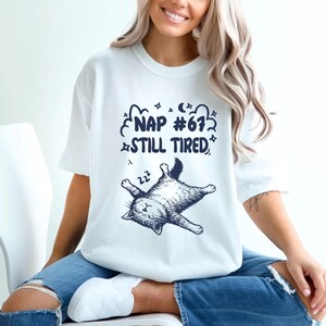 Op de afbeelding: Wit T-shirt met een marineblauwe afbeelding van een slapende kat en de tekst "NAP #67 STILL TIRED". De kat ligt op zijn rug met zijn poten in de lucht. Het shirt heeft een casual stijl.