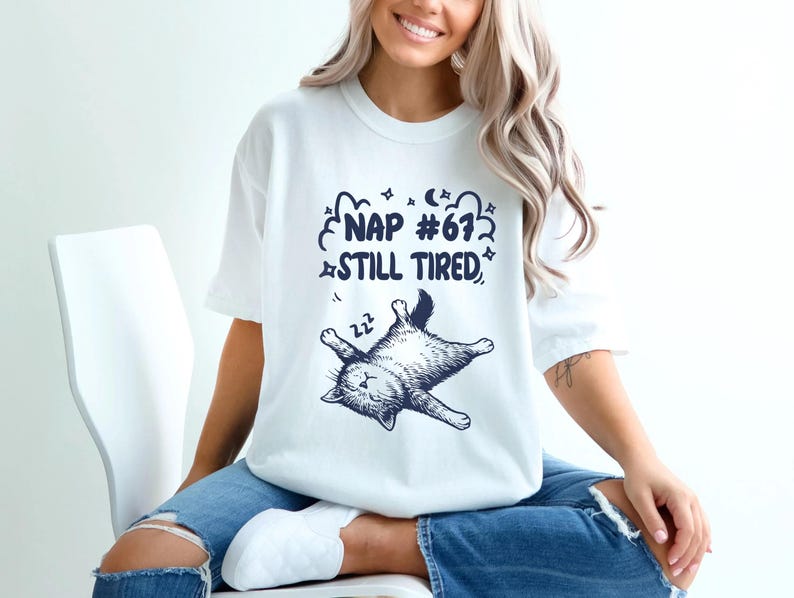 Op de afbeelding: Wit T-shirt met een marineblauwe afbeelding van een slapende kat en de tekst "NAP #67 STILL TIRED". De kat ligt op zijn rug met zijn poten in de lucht. Het shirt heeft een casual stijl.