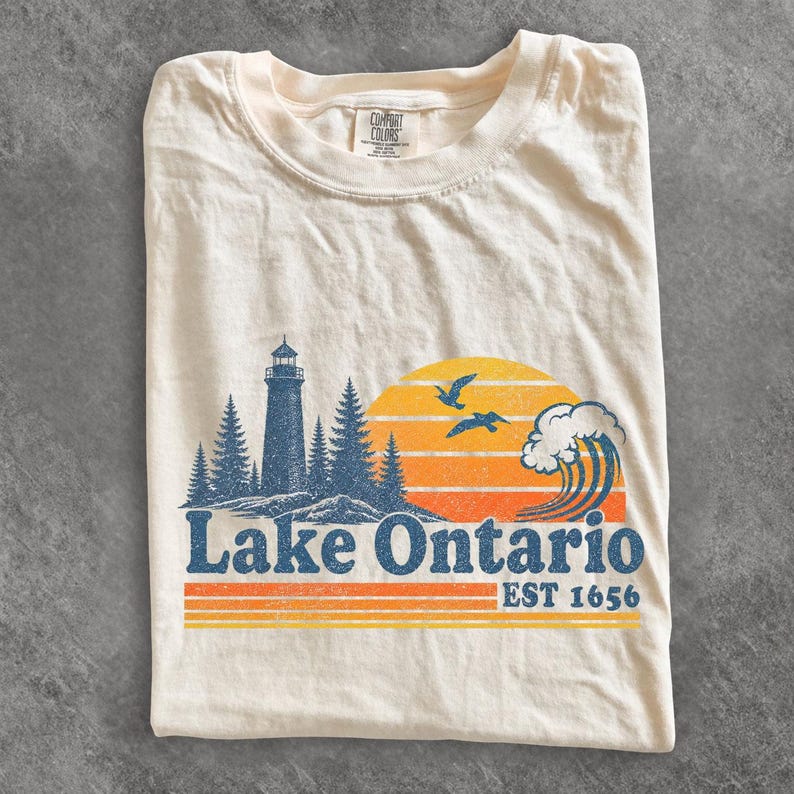 May include: A cream-colored short-sleeve t-shirt featuring a graphic of a lighthouse, pine trees, a large wave, and two birds against a sunset. The text reads Lake Ontario EST 1656 in blue letters with orange and yellow horizontal stripes below.