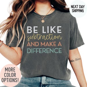 May include: Dark gray t-shirt with the text "BE LIKE subtraction AND MAKE A DIFFERENCE" in white, yellow, coral, and teal. The shirt has a crew neck and short sleeves. Additional text on the shirt says "MORE COLOR OPTIONS!" and "NEXT DAY SHIPPING".