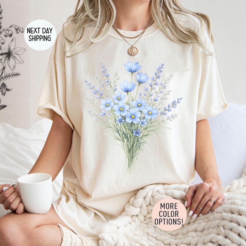 May include: Cream-coloured t-shirt with a watercolour bouquet of blue flowers and greenery. The shirt has short sleeves and a relaxed fit. The text "MORE COLOR OPTIONS!" is visible on the bottom right.