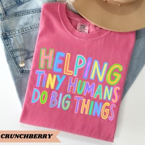 Op de afbeelding: Een roze T-shirt met korte mouwen en de tekst HELPING TINY HUMANS DO BIG THINGS in een meerkleurig lettertype met omlijning. Het shirt heeft een Comfort Colors-label bij de binnenhals en ligt op een lichtblauwe spijkerbroek.