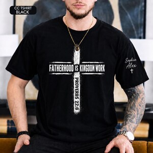 May include: Black t-shirt with a white cross design. The cross features the text "FATHERHOOD IS KINGDOM WORK" and "PROVERBS 22:6". The upper left corner has the text "CC TSHIRT BLACK". The right sleeve has the name "Sophia Alex" and a cross.