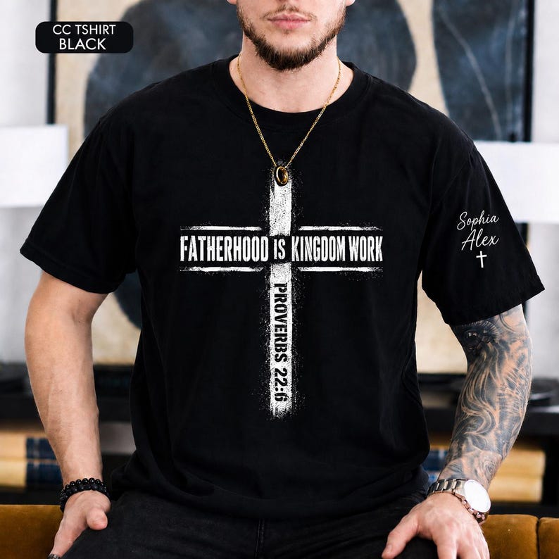 May include: Black t-shirt with a white cross design. The cross features the text "FATHERHOOD IS KINGDOM WORK" and "PROVERBS 22:6". The upper left corner has the text "CC TSHIRT BLACK". The right sleeve has the name "Sophia Alex" and a cross.