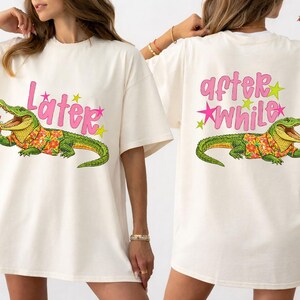 May include: Ivory-colored t-shirt featuring a cartoon alligator wearing a Hawaiian shirt and holding a drink. The front of the shirt has the word "Later" in pink script with star accents. The back of the shirt has the words "after while".