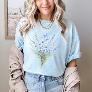 May include: Pale blue t-shirt with a watercolour floral design of blue flowers and green leaves. The shirt is made of soft material and is worn with blue jeans and a beige jacket. The style is casual and feminine.