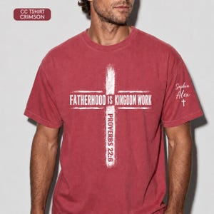 May include: Crimson red t-shirt with a white cross design. The cross features the text "FATHERHOOD IS KINGDOM WORK" and "PROVERBS 22:6". The shirt also has the name "Sophia Alex" and a cross on the sleeve.