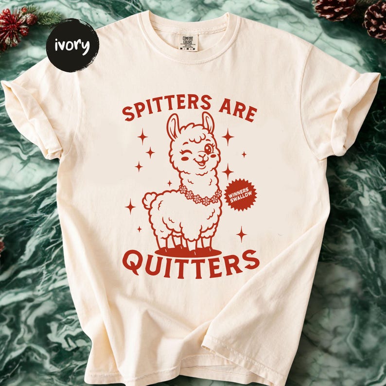 May include: Cream-coloured t-shirt featuring a red graphic of a smiling llama adorned with a floral necklace. The text "SPITTERS ARE QUITTERS" is above and below the llama. A small badge reads "WINNERS SWALLOW".