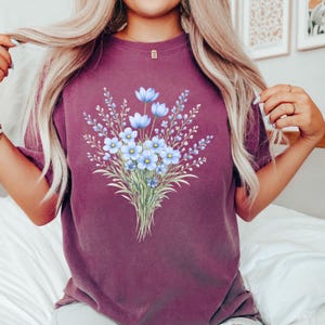 May include: A maroon t-shirt with a floral bouquet design. The bouquet features light blue flowers with green stems and leaves. The shirt is a solid maroon colour, with a crew neck and short sleeves.