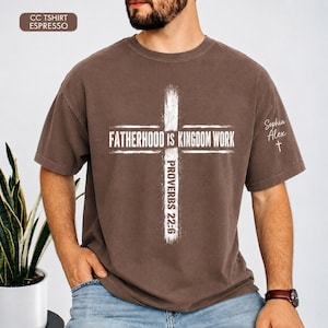 May include: Espresso-colored t-shirt with a white cross design. The text "FATHERHOOD IS KINGDOM WORK" is centered within the cross, with "PROVERBS 22:6" below. The sleeve features the name "Sophia Alex" and a small cross.