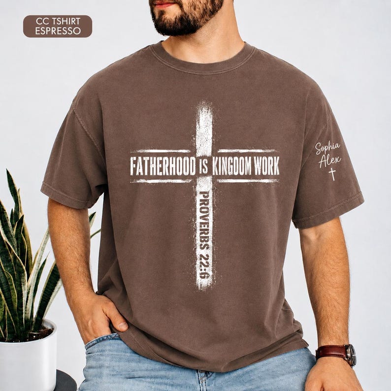 May include: Espresso-colored t-shirt with a white cross design. The text "FATHERHOOD IS KINGDOM WORK" is centered within the cross, with "PROVERBS 22:6" below. The sleeve features the name "Sophia Alex" and a small cross.