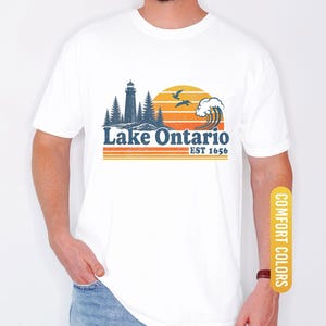 May include: A white t-shirt featuring a graphic print of a lighthouse, pine trees, a crashing wave, and two birds against a sunset. The text reads Lake Ontario EST 1656 in blue and orange. The design includes horizontal orange stripes below the text.