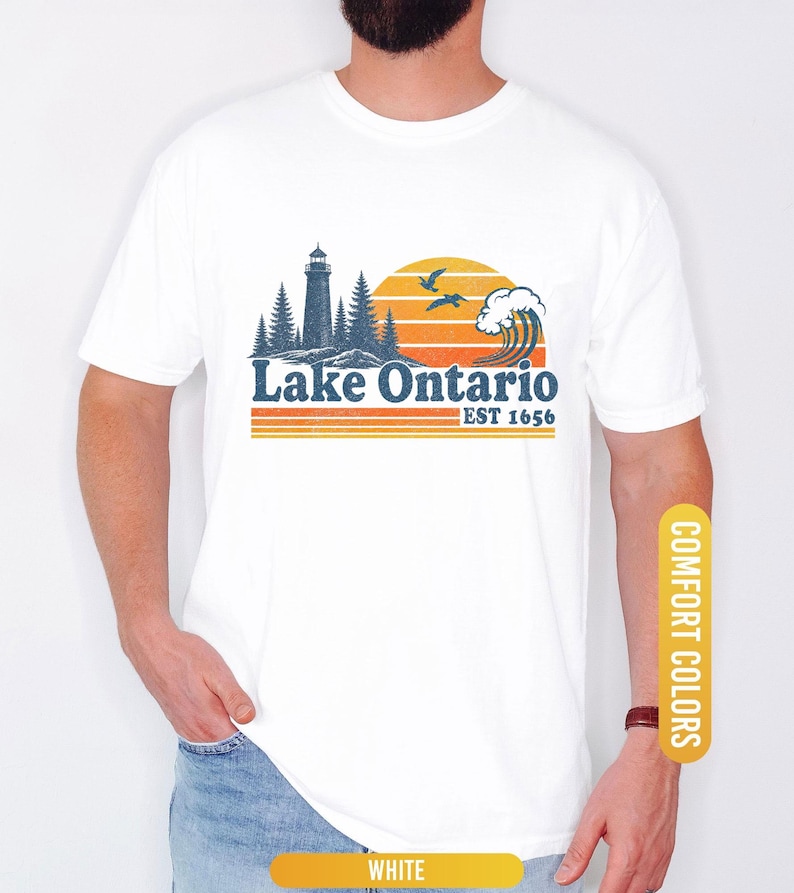 May include: A white t-shirt featuring a graphic print of a lighthouse, pine trees, a crashing wave, and two birds against a sunset. The text reads Lake Ontario EST 1656 in blue and orange. The design includes horizontal orange stripes below the text.