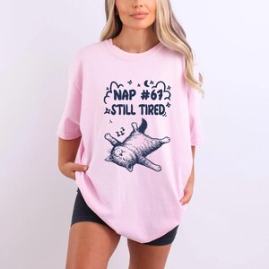 Op de afbeelding: Lichtroze T-shirt met een slapende kat en de tekst "NAP #67 STILL TIRED" in marineblauw. Het T-shirt heeft een losse pasvorm.