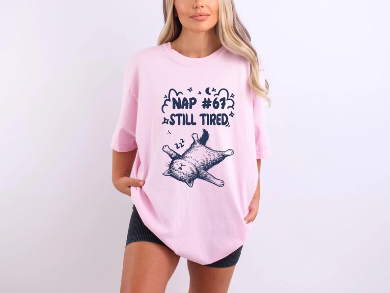 Op de afbeelding: Lichtroze T-shirt met een slapende kat en de tekst "NAP #67 STILL TIRED" in marineblauw. Het T-shirt heeft een losse pasvorm.