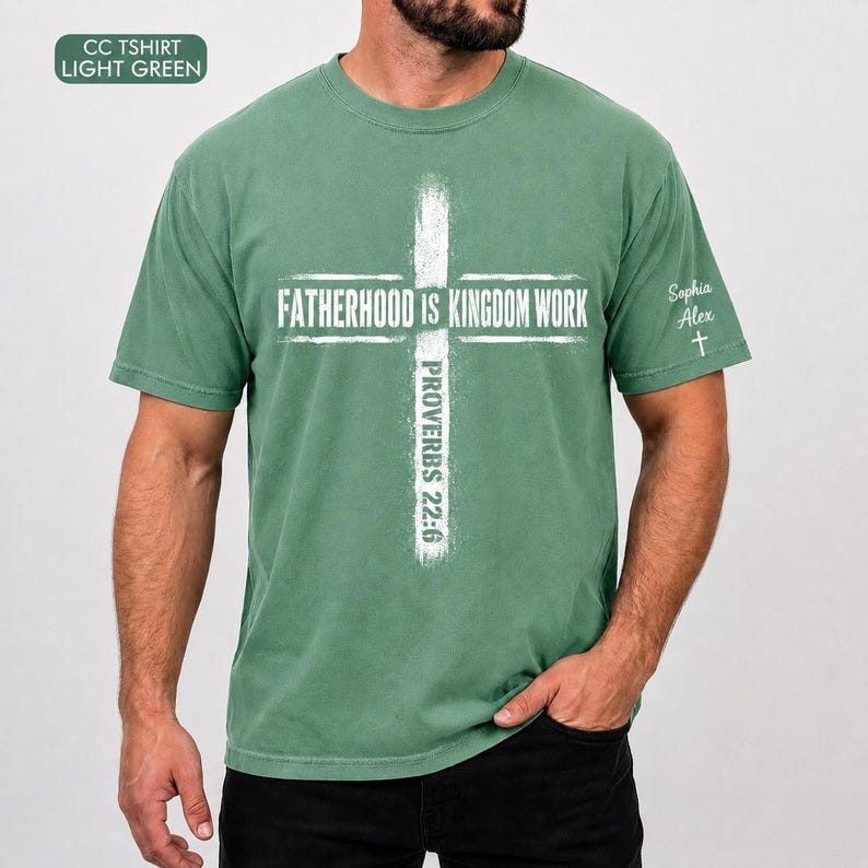May include: Light green t-shirt with the text "FATHERHOOD IS KINGDOM WORK" in white, forming a cross shape. The text "PROVERBS 22:6" is printed vertically. The shirt also has the text "Sophia Alex" and a cross symbol on the sleeve.