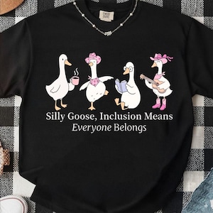以下が含まれることがあります： 黒いスウェットシャツ。4羽の白いガチョウの気まぐれなイラストが特徴です。各ガチョウは、コーヒーカップ、カウボーイハット、メガネ、ギターなど、ユニークなアクセサリーを身に着けています。「Silly Goose, Inclusion Means Everyone Belongs」というテキストが下に印刷されています。
