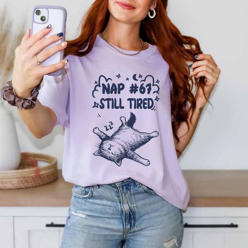 Op de afbeelding: Een lichtpaars t-shirt met de tekst "NAP #672 STILL TIRED" en een slapende kattenafbeelding. Het shirt is gemaakt van een zachte stof en is geschikt voor vrijetijdskleding. Het ontwerp is gedrukt met zwarte inkt.