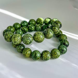 May include: Two green beaded bracelets. The beads are round and have a speckled pattern in shades of green. The bracelets are resting on a white surface.