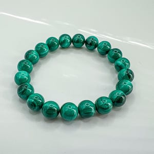 May include: A bracelet featuring round, polished beads of malachite. The beads are a vibrant green with darker green swirling patterns. The bracelet is strung on an elastic band, creating a circular shape.