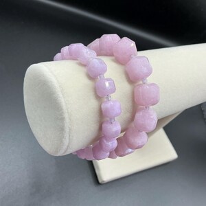 May include: Two light purple beaded bracelets. The bracelets are displayed on a white, cylindrical stand. The beads are faceted and have a square shape. The background is black.