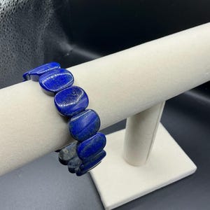 May include: A dark blue gemstone bracelet with oval-shaped beads. The bracelet is displayed on a white jewelry stand against a black background. The beads have a polished finish and vary slightly in color.