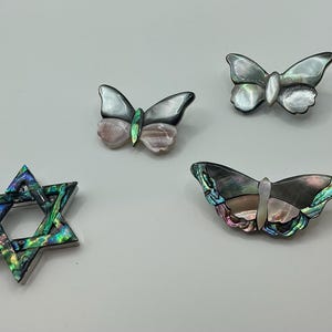 May include: Four decorative brooches. Three butterfly-shaped brooches with iridescent wings in shades of gray, pink, and green. A Star of David brooch with a similar iridescent finish. All brooches are set against a white background.