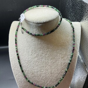 May include: A necklace and bracelet set featuring small, square-cut beads in shades of green, black, and red. The jewelry is displayed on a white bust against a dark background, showcasing the natural gemstone colors and design.