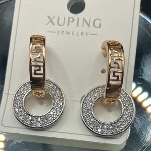 May include: A pair of gold-toned hoop earrings with a geometric design and a circular pendant encrusted with small, sparkling crystals. The earrings are displayed on a white card with the text "XUPING - JEWELRY".