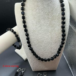 May include: A black beaded necklace, bracelet, and two pairs of earrings are displayed on a white mannequin. The jewelry features square-cut beads. The earrings are labeled "3 stone earring". The background is a mix of gray and silver.