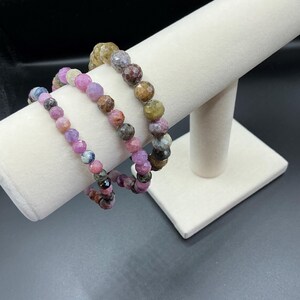 May include: Three beaded bracelets with faceted gemstones in shades of pink, purple, brown, and green are displayed on a white, cylindrical jewelry stand. The bracelets are strung on elastic and appear to be handmade.