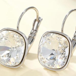 May include: A pair of silver-toned leverback earrings. Each earring features a large, square-cut, clear crystal stone set in a silver frame. The crystals have multiple facets, reflecting light. The earrings are displayed on a light beige background.