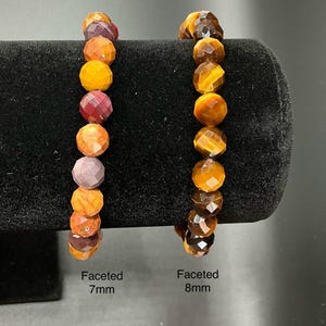 May include: Two beaded bracelets with faceted gemstones. One bracelet features 7mm beads in shades of orange, red, and purple. The other bracelet has 8mm tiger's eye beads. Both bracelets are displayed on a black surface.