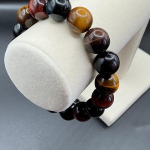May include: A beaded bracelet featuring large, polished beads in shades of brown, black, and red. The bracelet is displayed on a white stand, showcasing the variety of colors and textures of the gemstones.